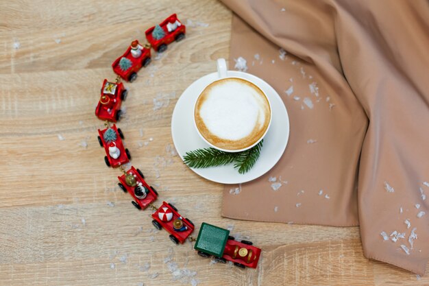 Delicious fresh festive morning cappuccino coffee in a ceramic white cup on the wooden table with decorative christmas train, red ornamentals, fireflies and spruce branches