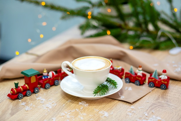 装飾的なクリスマストレイン、赤い装飾品、ホタル、スプルースの枝と木製のテーブルの上の白いセラミックカップでおいしい新鮮なお祝いの朝カプチーノコーヒー