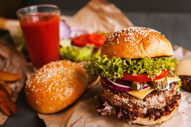 Delicious fresh homemade burger on a wooden table. next to the component to burger, wooden trays, fried potatoes and chili pepper. A glass of tomato juice