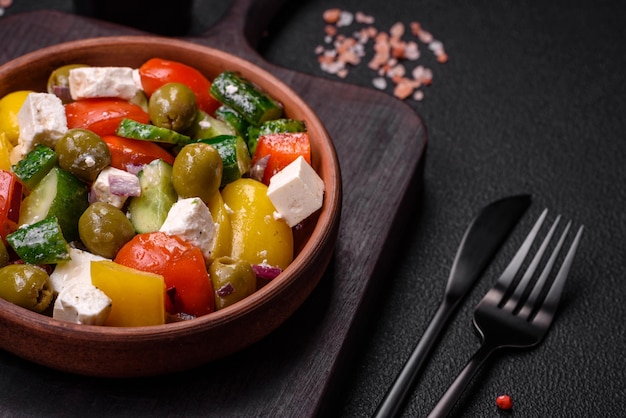 Вкусный свежий сочный греческий салат с сыром фета, оливками, помидорами, огурцом, перцем, солью и специями на темном бетонном фоне.