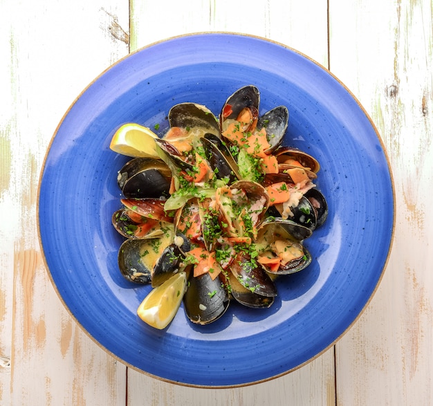 Delicious freshly cooked black mussels in tomato sauce in a vintage saucepan with spoon next to slice of lemon on a wooden background
