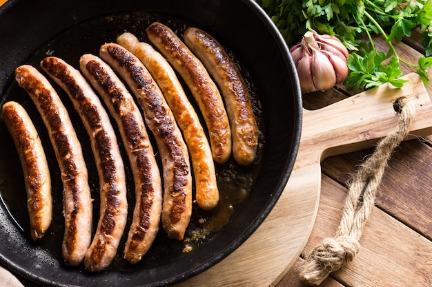 Delicious fried sausages with golden crust in iron cast pan, fresh parsley garlic on wood table