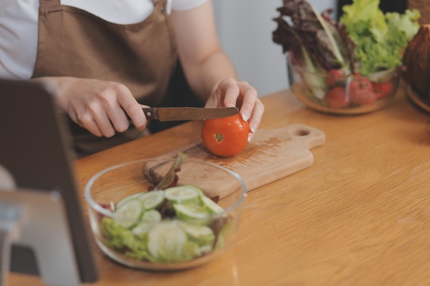 На столе вкусные фрукты и овощи, а женщина готовит. Домохозяйка режет зеленые огурцы на деревянной доске для приготовления свежего салата на кухне.