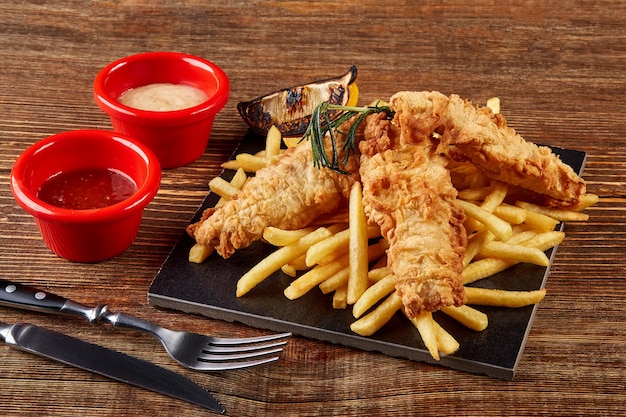 Delicious golden batter deep fried fish fillets and French fries, served on black board on wooden worktop, view from above.