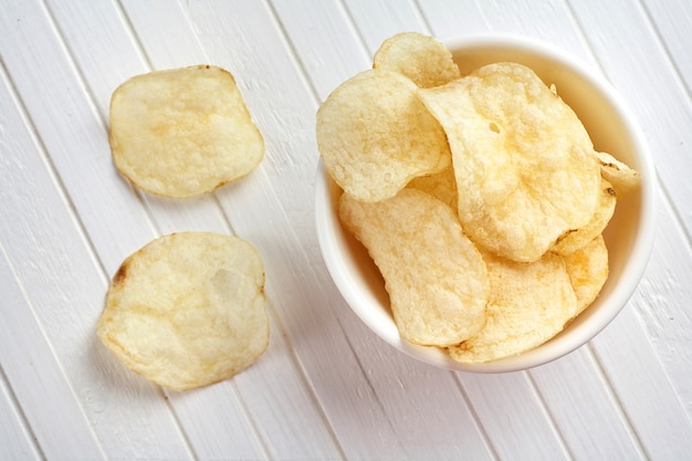 Delicious golden potato chips in a white bowl on a white wooden background.