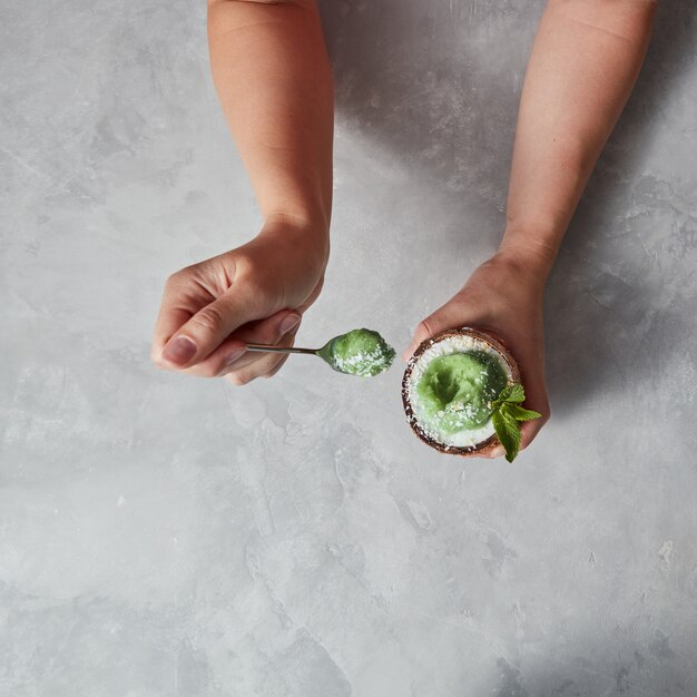 Delicious green dessert, ice cream in a coconut shell in the woman hand with spoon on a gray concrete table with copy space. Top view.