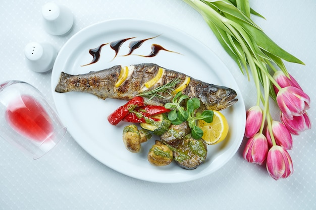 Delicious grilled seabass with lemonnd a side dish of grilled vegetables (mushrooms, tomatoes, bell peppers) on white plate.