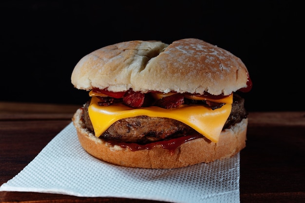 Delizioso hamburger con pancetta e formaggio cheddar su pane fatto in casa con semi e ketchup su una superficie di legno e sfondo nero.