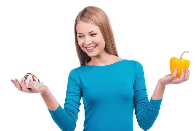 Delicious and harmful or helpful and tasty? Beautiful young woman holding a salad pepper in one hand and a cake in another  while standing against white background