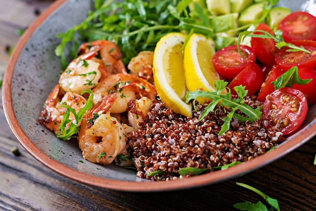 Delicious healthy Buddha bowl with shrimps, tomato, avocado, quinoa, lemon and arugula on the wooden table. Healthy food.