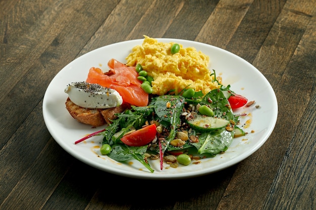 Delicious and hearty breakfast - scrambled eggs and toast with cream cheese and salmon, mix salad in a white plate on a wooden surface