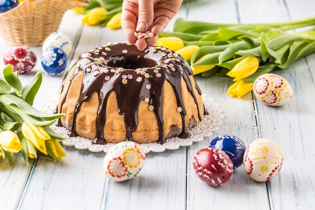 Вкусный праздничный словацкий и чешский торт бабовка с шоколадной глазурью. Женские руки украшают торт. Пасхальные украшения - весенние тюльпаны и яйца.