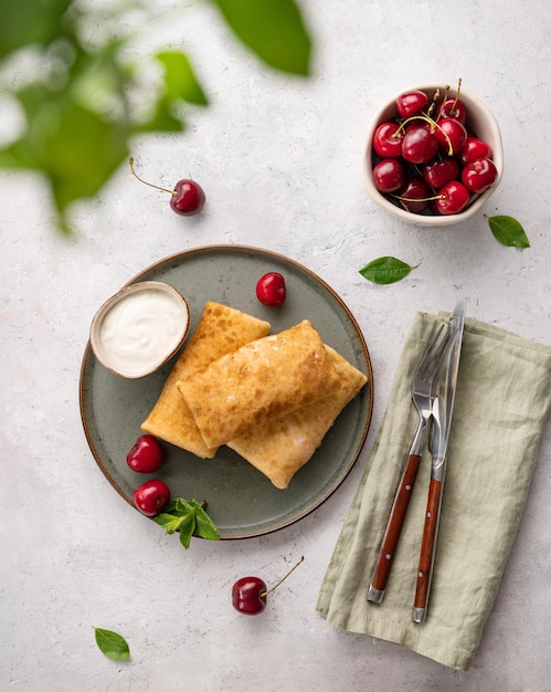 Delicious homemade pancakes or crepes with fresh cherries and sour cream on a dark plate on a light background Top view