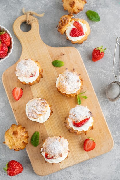 Delicious homemade small cakes profiterole choux pastry with custard, strawberry and icing powder on a gray concrete background. Copy space.