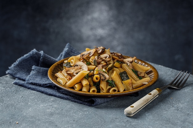 Photo delicious italian dish, penne pasta with spanach, zucchini and roasted mushrooms with garlic on the blue marble textured table with empty space for text. integral pasta and whole champignon mushrooms