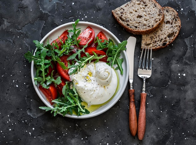 Вкусный салат в итальянском стиле, свежая руккола буррата и помидоры на темном фоне, вид сверху