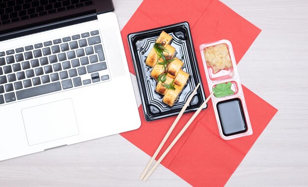 Delicious Japanese rolls with salmon avocado cucumber cheese in takeout packaging with wood sticks soy sauce and laptop on office table Selective focus Asian delivery food concept