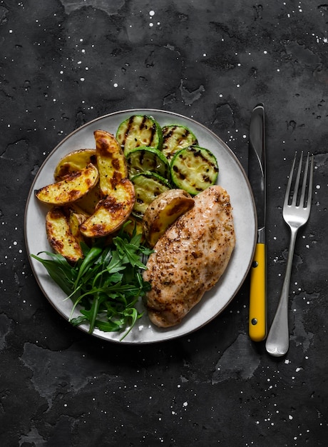 Delicious lunch baked potatoes chicken breast grilled zucchini\
and arugula spinach salad on a dark background the view is\
super