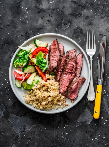 Delicious lunch bulgur lettuce cucumber tomato salad and steak on a dark background top view