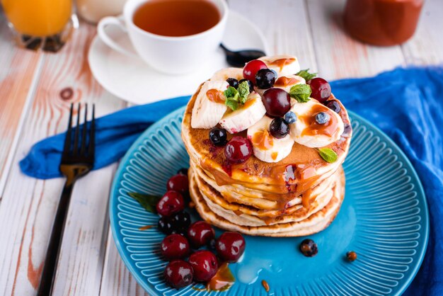 Delicious and lush pancakes with fresh fruits and berries, poured with salted caramel. Mint, tea, orange juice, milk on the table.