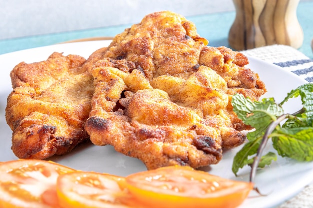 Photo delicious milanese chicken breaded chicken with breadcrumbs and egg a typical brazilian dish on a white plate with tomatoes lemon on striped white tablecloth and blue marble