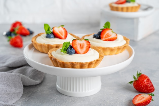 Delicious mini tarts with fresh berries and custard on a white plate on a gray concrete background