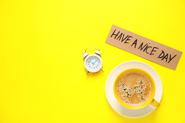 Delicious morning coffee alarm clock and card with HAVE A NICE DAY wish on yellow background flat lay Space for text