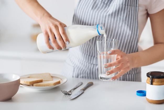 맛있고 영양가 있습니다. 밝은 부엌에 서서 우유를 유리에 모으는 동안 아침 식사를 준비하는 조심스러운 귀여운 사랑하는 여자