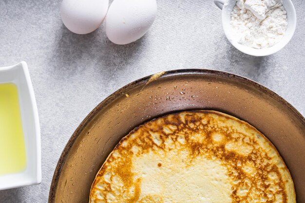 Delicious pancakes in a frying pan on a white table with wheat eggs olive oil tomato sauce and a checkered tablecloth in the background