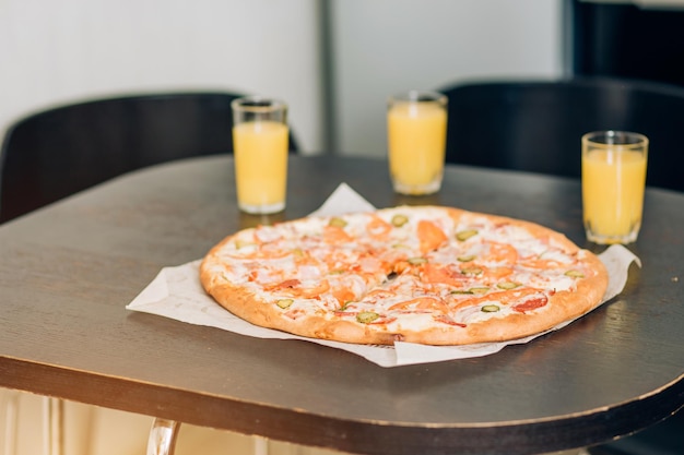 Delicious pizza and three glasses of orange juice on table. Cropped shot of man pouring drinks into glass. Selective focus. Food concept.