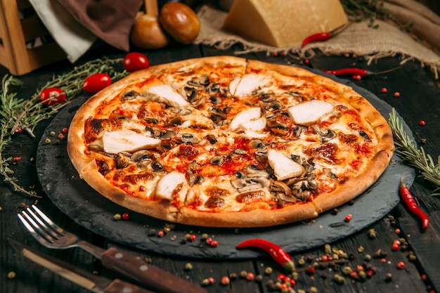 Delicious pizza with mushrooms and chicken on the black board on the dark wooden table with herbs and tomatoes