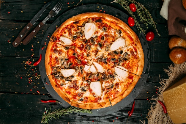 Delicious pizza with mushrooms and chicken on the black board on the dark wooden table with herbs and tomatoes