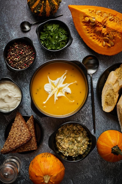 Delicious pumpkin soup with cream seeds bread and fresh herbs in elegant ceramic black bowl on dark stone background Top view flat lay Autumn food concept