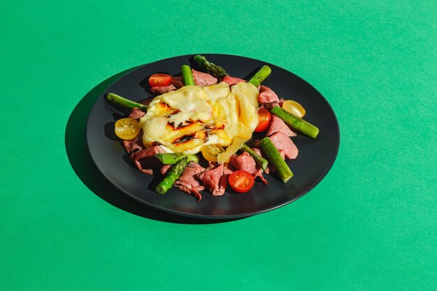 Delicious raclette cheese served over Roast Beef with Tomatoes and Asparagus dish on green background Hard light deep shadow