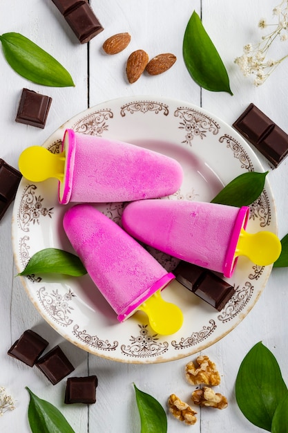 Delicious and refreshing ice cream lollies in a vintage plate on a white wooden table covered with pieces of chocolate, nuts, almonds and leaves from nature