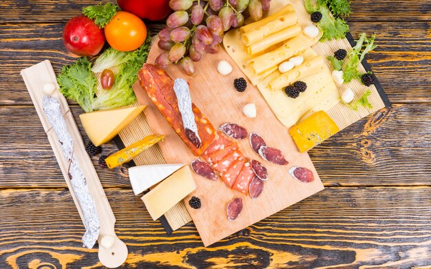 Delicious selection of different cheeses and spicy sausage displayed on a cheeseboard with grapes garnished with fresh blackberries, olives and bell peppers, overhead view