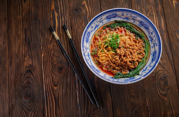 Delicious and spicy homemade sichuan rice noodle in traditional blue bowl with black chopsticks on wooden table, asian food concept copy space for menu design