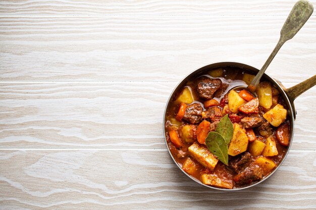 Photo delicious stew with meat, potatoes, carrot and gravy in rustic copper pot on white wooden background from above. traditional winter and autumn dish beef and vegetables ragout in stewpot, copy space