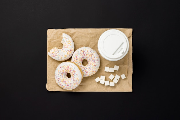 Delicious, sweet, fresh donuts, paper cup of coffee, paper packaging bag for takeaway food, black background. Concept of breakfast, fast food, coffee shop, bakery, lunch. Flat lay, top view.