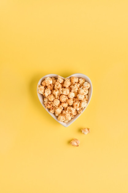 Delicious sweet popcorn with caramel in white ceramic heart plate, isolated on trend color yellow table.