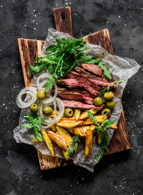 Delicious tapas board medium rare beef steak french fries green salad olives pickled onions on a dark background top view