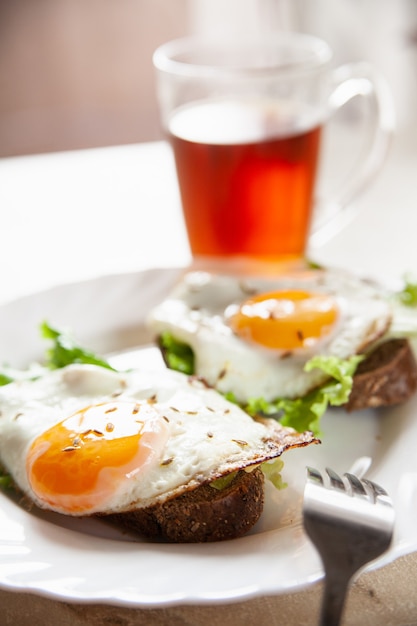 Delicious toasts with fried eggs and lettuce leaves for breakfast placed on table near cup of hot tea in morning