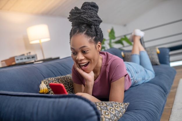 Delight, emotion. Enthusiastic surprised young african american woman with black hair looking at smartphone lying on sofa at home