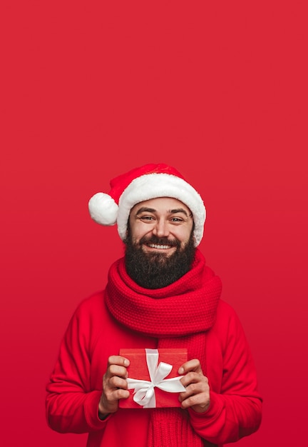 Delighted adult bearded male in Santa hat and scarf holding small gift