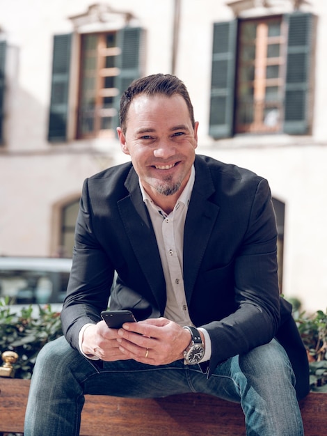 Delighted adult male manager with dark hair and bear in stylish smart casual outfit smiling happily and looking at camera while using mobile phone sitting on wooden bench on street