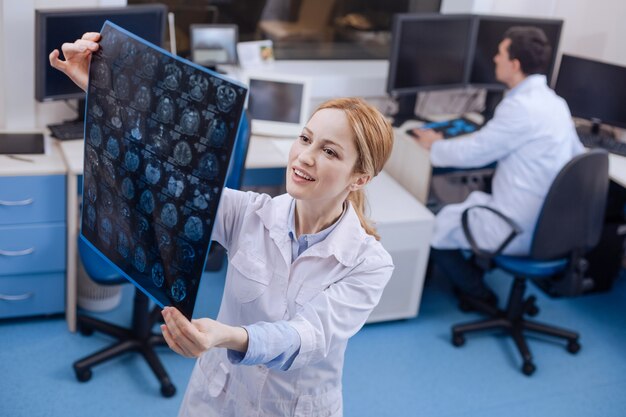 Delighted attractive female doctor standing in her lab and looking at the x ray images while being in a positive mood