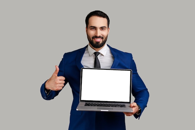 Delighted bearded man working on laptop with smile and showing thumb up looking at camera