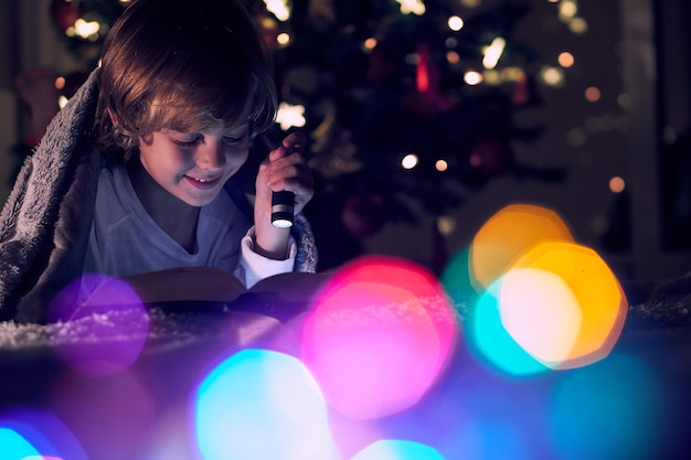 Delighted boy reading book with flashlight while lying under blanket in dark room with colorful bokeh and bright glowing lights