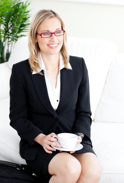 Delighted businesswoman wearing glasses drinking coffee sitting on a sofa