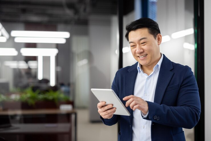 Photo delighted chinese head of department wearing formal suit and using digital tablet with wide smile happy brunette man enjoying modern technology and new opportunities for remote work copy space